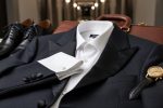 Bond-inspired tailored menswear ensemble with dark tuxedo jacket, white dress shirt, vintage gold watch, and polished leather shoes arranged on a leather briefcase in a luxury setting.