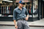 A man modeling an early-2000s-inspired outfit with a trucker hat, popped-collar shirt, cargo shorts, and a studded belt in an urban streetwear setting.